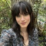 A person with long dark hair and bangs takes a selfie outdoors in front of trees and greenery, wearing a patterned top.