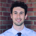 A man with short dark hair and light facial hair, wearing a light blue shirt and dark tie, stands in front of a brick wall, smiling.